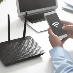Ręce trzymają smartfon z ikoną Wi-Fi, obok router i laptop. Dowiedz się, jak sprawdzić na jakiej częstotliwości pracuje Wi-Fi.