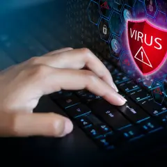 Dłoń naciska klawisz Enter na klawiaturze, symbolizując walkę z zagrożeniem. Ikona tarczy z napisem "VIRUS" i wykrzyknikiem sugeruje, jak usunąć wirusa z komputera.