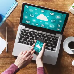 Ręce trzymają smartfon, który pokazuje ikony chmury. Obok laptop z podobnym ekranem. To inspiracja, jak połączyć komputer z internetem z telefonu.