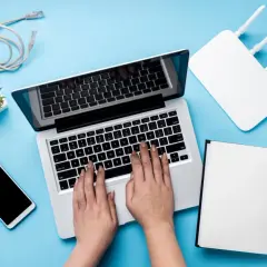 Ręce na klawiaturze laptopa, obok router wifi, telefon i kabel. Szukasz sposobu, jak zwiększyć zasięg wifi w laptopie?