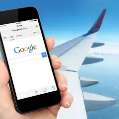 Dłoń trzyma smartfon z otwartą wyszukiwarką Google, na tle skrzydła samolotu i nieba. Czy w samolocie można mieć internet? Tak, można!