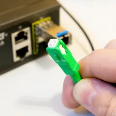 Dłoń trzyma zieloną wtyczkę światłowodową, gotową do podłączenia do portu w przełączniku Ethernet.