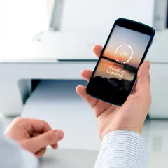 Dłoń trzyma smartfon z napisem "Sending to printer" i wskaźnikiem postępu 37%. W tle biała drukarka.