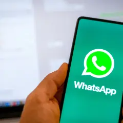 Dłoń trzyma smartfon z logo WhatsApp. W tle widać ekran laptopa z otwartym programem do komunikacji, sugerującym użycie WhatsApp na komputer.