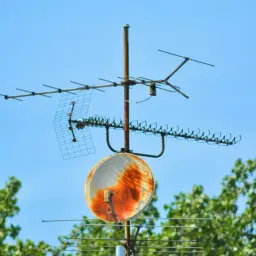 Zardzewiała antena satelitarna i telewizyjna pokojowa na tle błękitnego nieba, otoczona zielonymi drzewami.