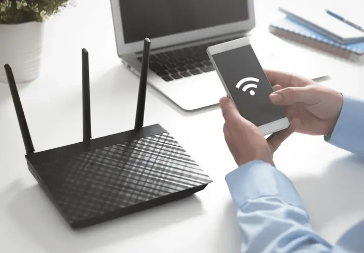 Ręce trzymają smartfon z ikoną Wi-Fi, obok router i laptop. Dowiedz się, jak sprawdzić na jakiej częstotliwości pracuje Wi-Fi.