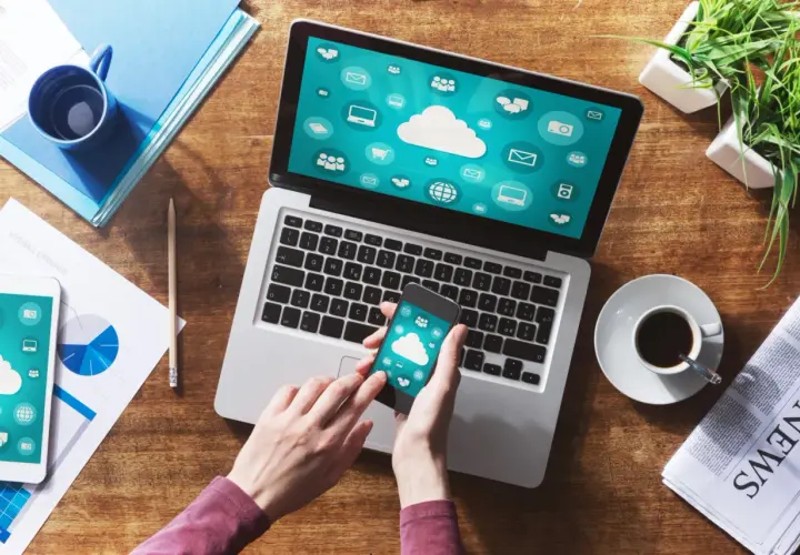 Ręce trzymają smartfon, który pokazuje ikony chmury. Obok laptop z podobnym ekranem. To inspiracja, jak połączyć komputer z internetem z telefonu.
