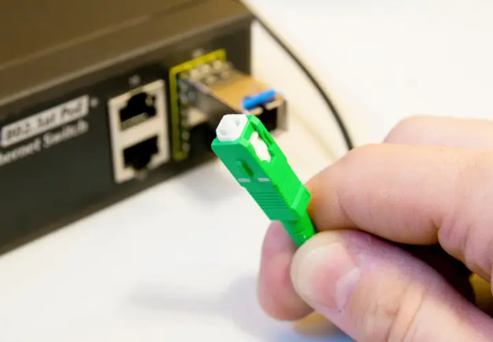 Dłoń trzyma zieloną wtyczkę światłowodową, gotową do podłączenia do portu w przełączniku Ethernet.