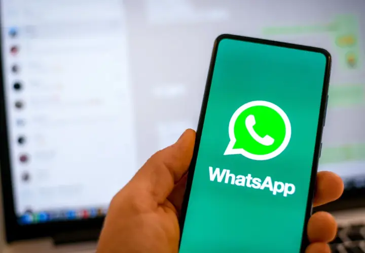 Dłoń trzyma smartfon z logo WhatsApp. W tle widać ekran laptopa z otwartym programem do komunikacji, sugerującym użycie WhatsApp na komputer.