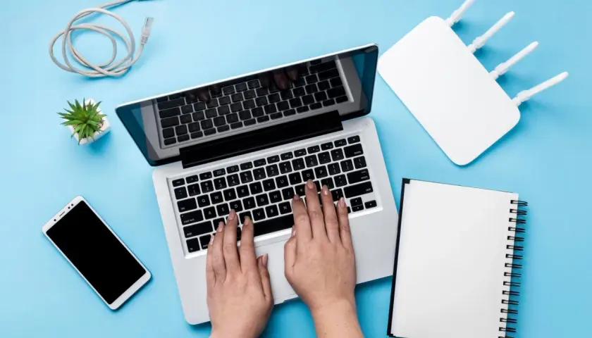 Ręce na klawiaturze laptopa, obok router wifi, telefon i kabel. Szukasz sposobu, jak zwiększyć zasięg wifi w laptopie?