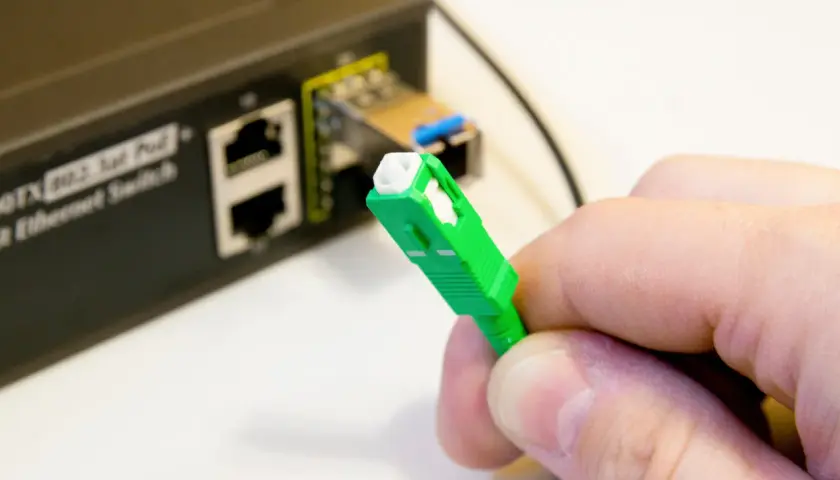 Dłoń trzyma zieloną wtyczkę światłowodową, gotową do podłączenia do portu w przełączniku Ethernet.