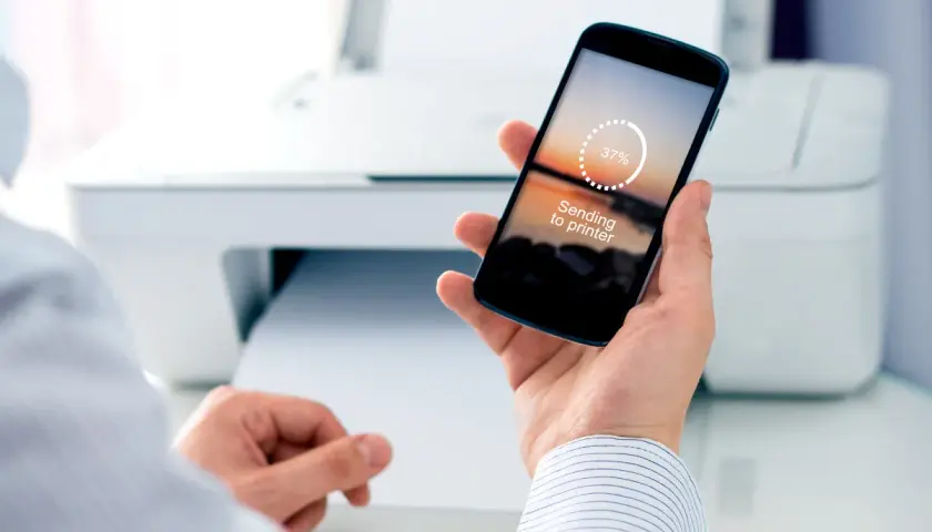 Dłoń trzyma smartfon z napisem "Sending to printer" i wskaźnikiem postępu 37%. W tle biała drukarka.