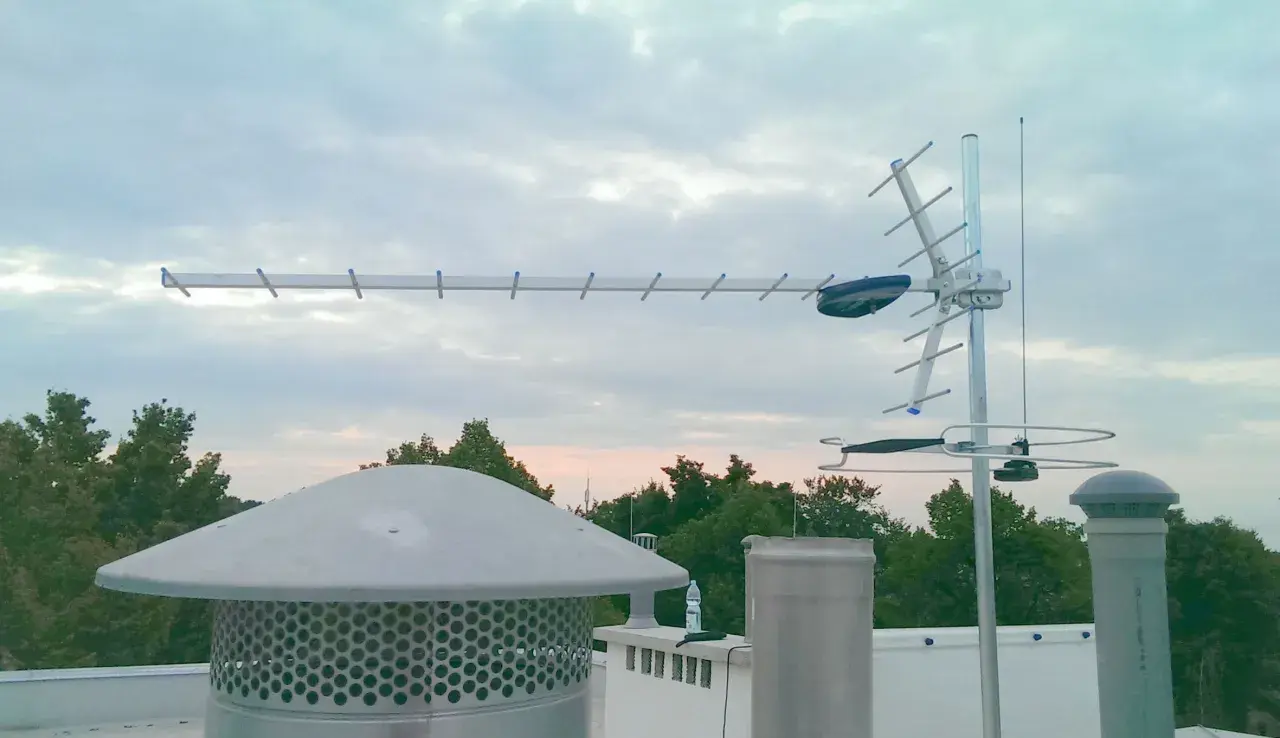 Antena telewizji naziemnej na dachu, na tle nieba i drzew.