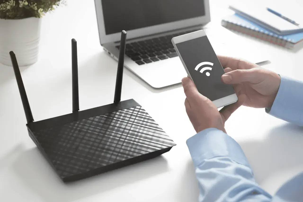 Ręce trzymają smartfon z ikoną Wi-Fi, obok router i laptop. Dowiedz się, jak sprawdzić na jakiej częstotliwości pracuje Wi-Fi.
