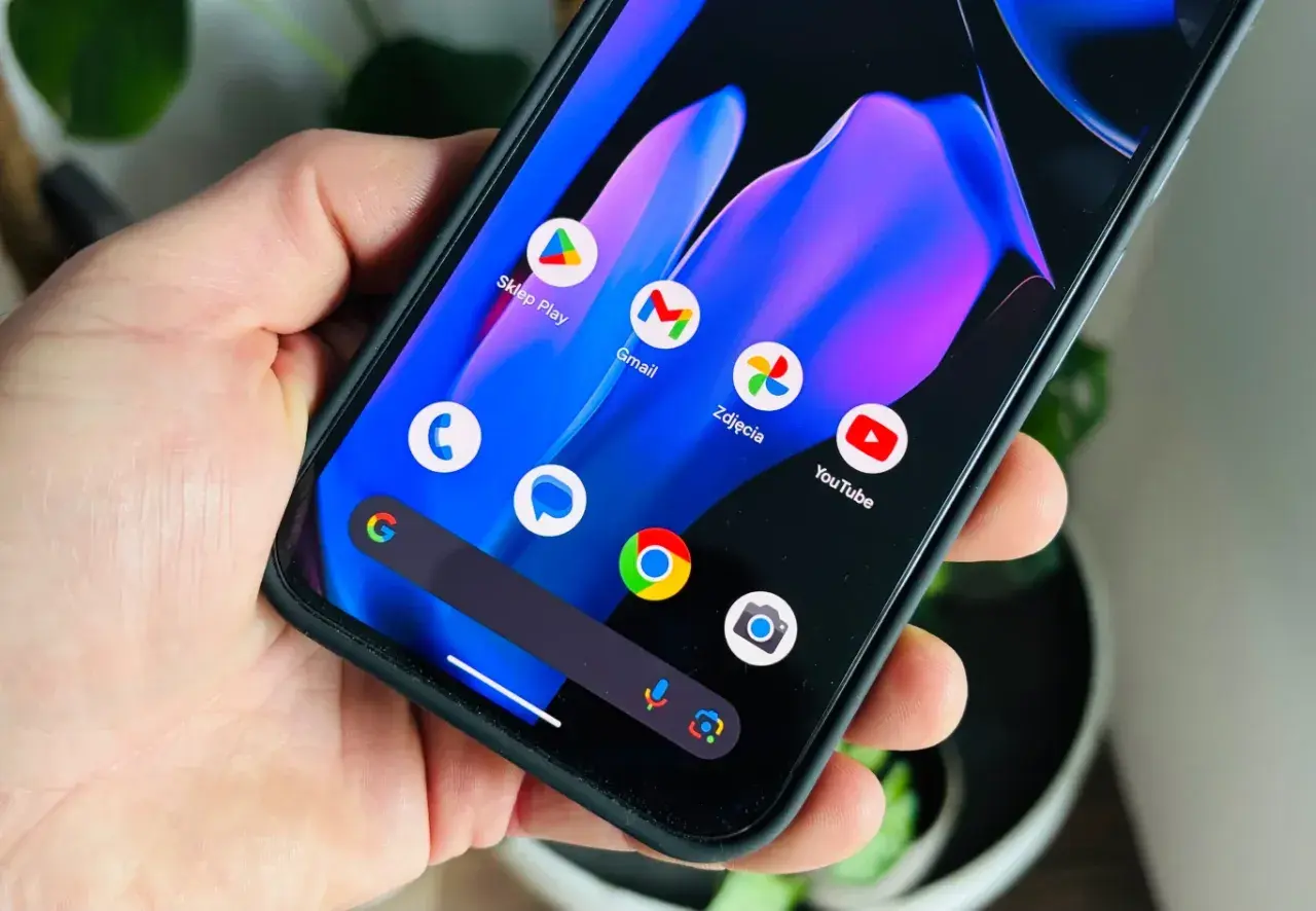 Dłoń trzyma smartfon z kolorową tapetą i ikonami aplikacji, w tym Sklep Play, Gmail, Zdjęcia i YouTube. Wygląda jak nowy **android 15**.