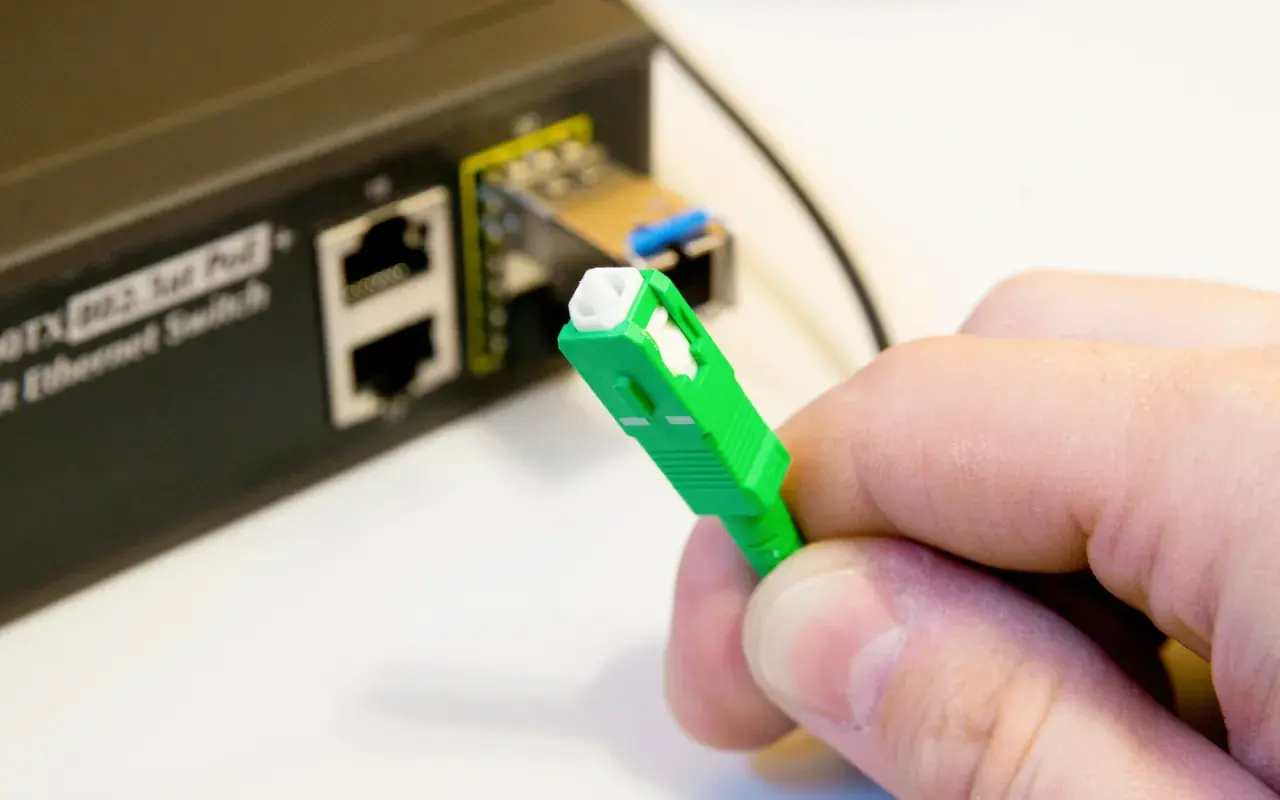 Dłoń trzyma zieloną wtyczkę światłowodową, gotową do podłączenia do portu w przełączniku Ethernet.