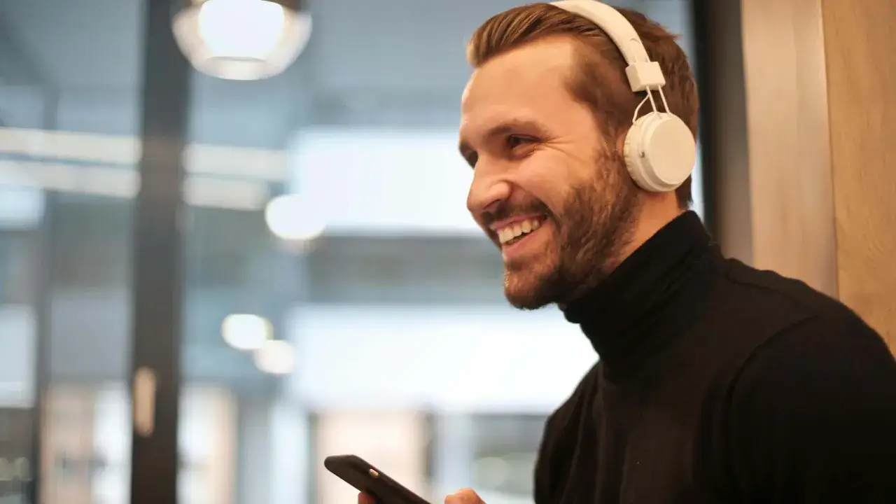 Uśmiechnięty mężczyzna w słuchawkach na uszach trzyma telefon, jakby sprawdzał, jak dodać słuchawki do telefonu.